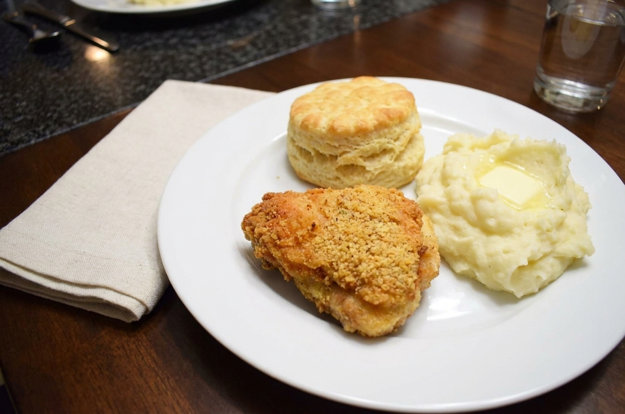 A dinner plate with crispy fried chicken, a flaky biscuit, and creamy mashed potatoes on a dining table with simple napkins under warm lighting