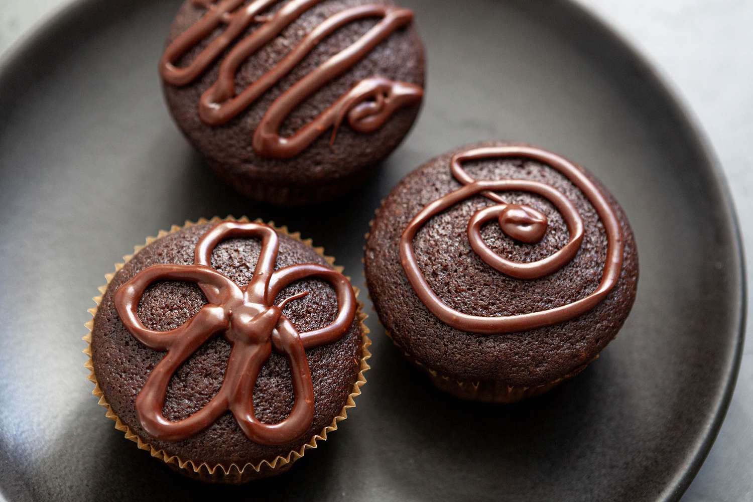 A dozen plain chocolate cupcakes with domed tops on a simple white plate on a warm kitchen table with natural window light