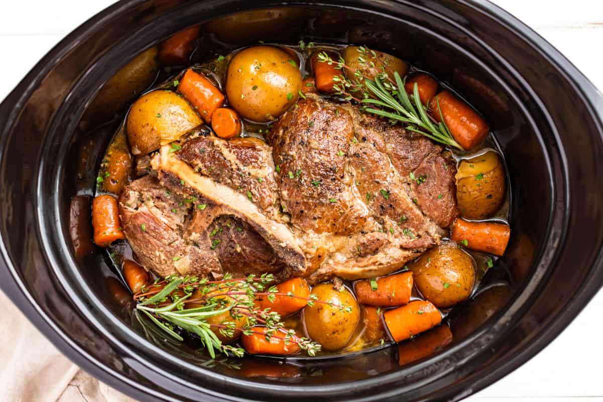 A fall-apart crock pot pork roast on a platter with carrots and baby potatoes, glossy herb gravy spooned over the top on a wooden kitchen table