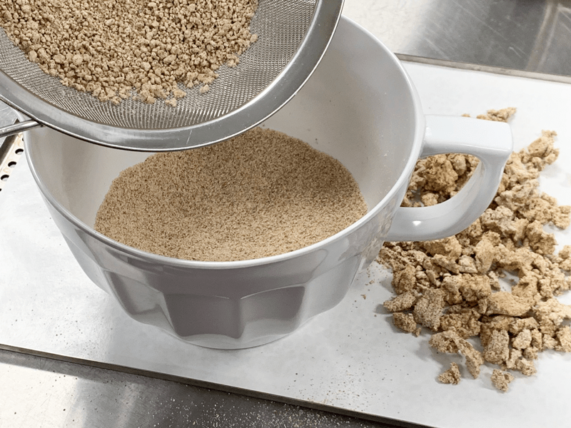 A fine-mesh sieve held over a bowl while almond flour and powdered sugar are sifted on a clean kitchen counter
