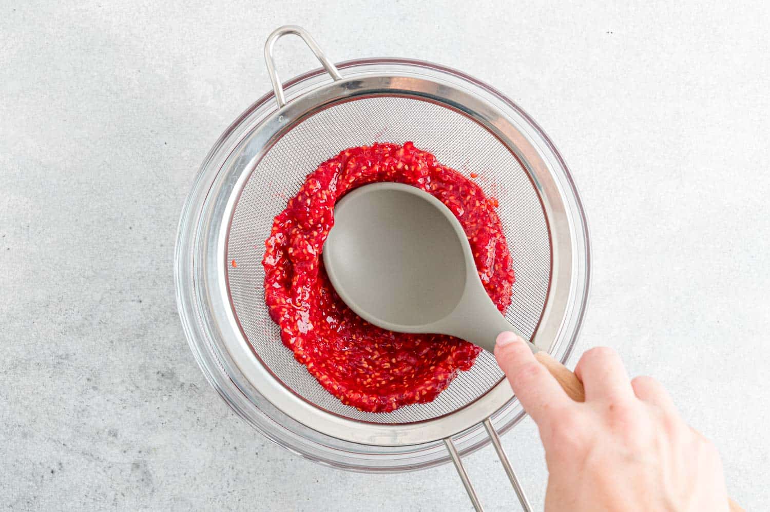 A fine-mesh strainer set over a bowl as bright pink raspberry vinaigrette is pressed through to remove seeds