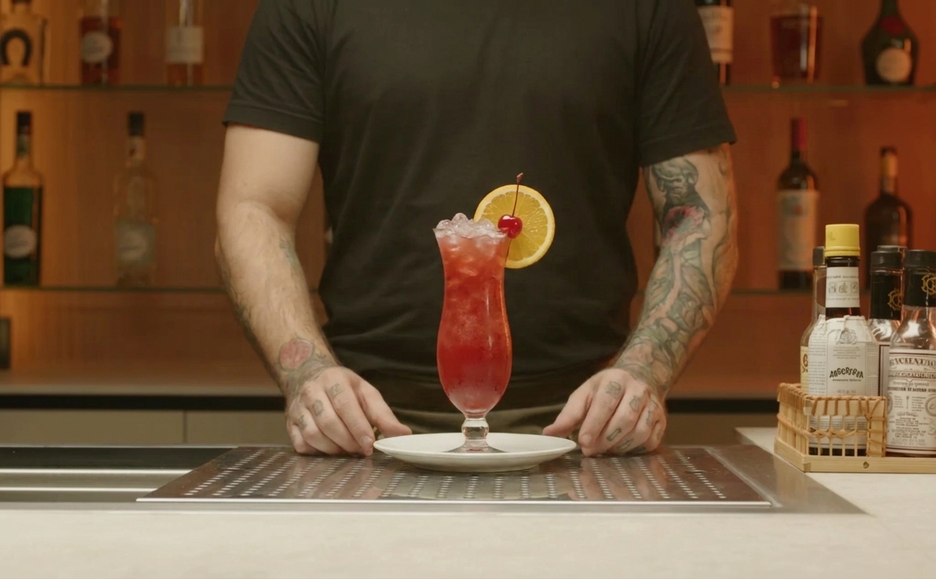 A finished Hurricane cocktail on a small plate with an orange slice and maraschino cherry garnish, condensation on the glass and crushed ice visible
