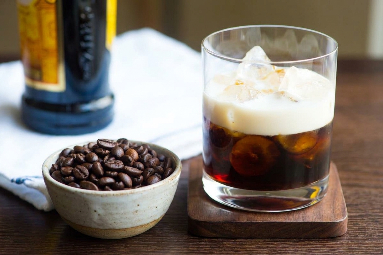 A finished White Russian cocktail in a rocks glass on a coaster with a small bowl of coffee beans nearby on a softly lit table