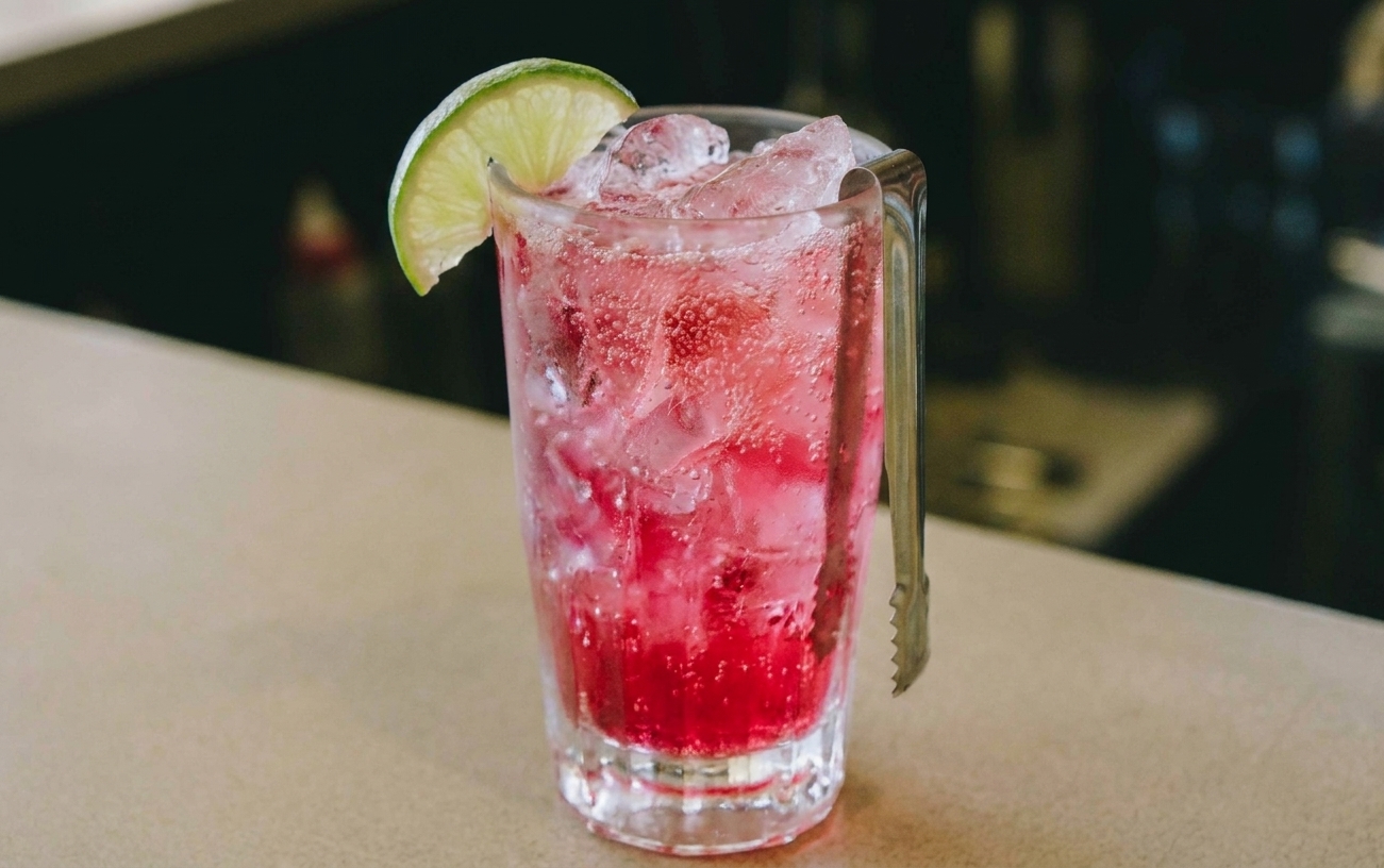A finished bright pink fizzy drink in a tall glass with a lime wedge garnish on a neutral tabletop