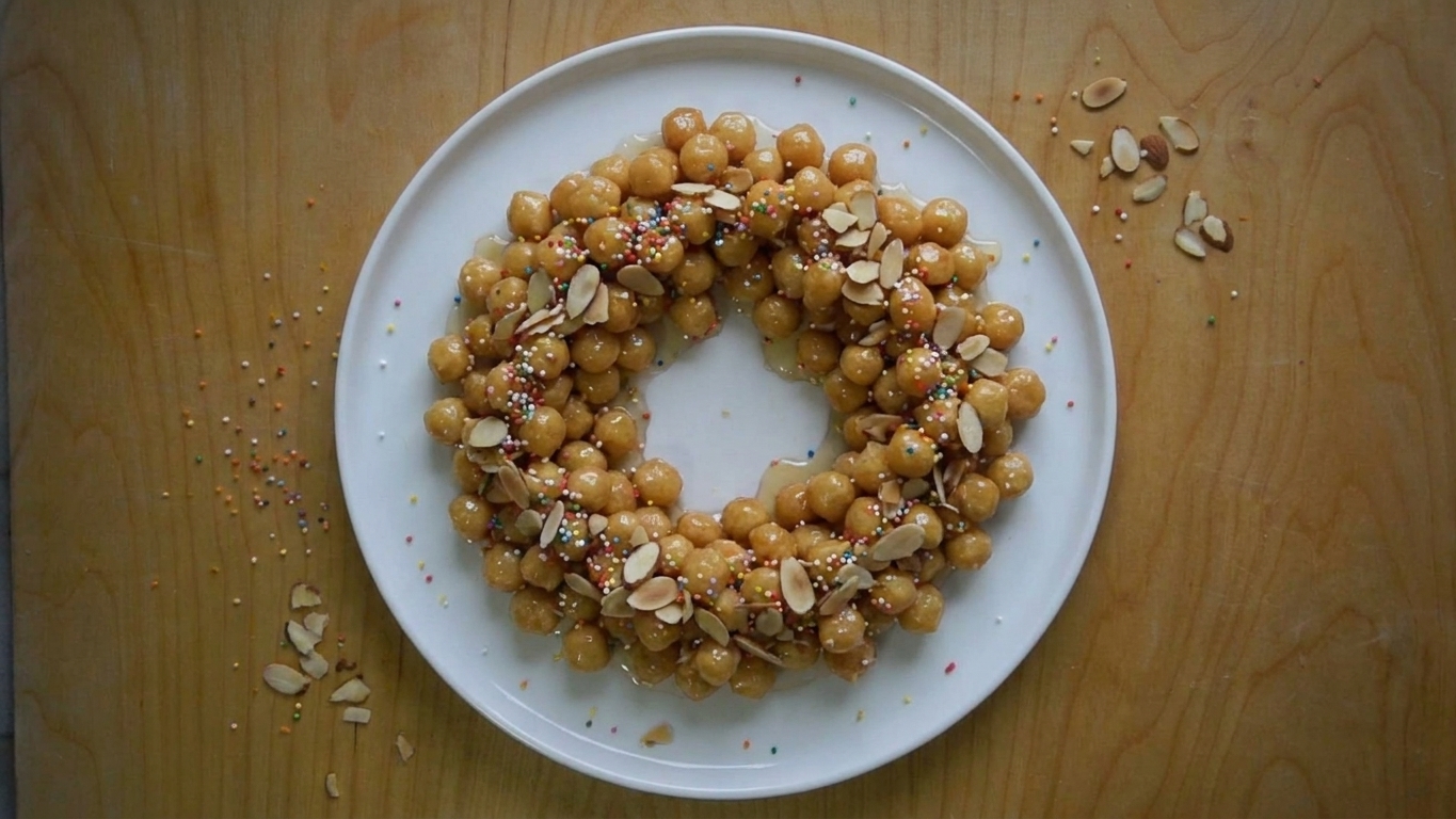 A finished platter of struffoli arranged in a wreath shape, shiny with honey glaze and topped with rainbow sprinkles and toasted almonds