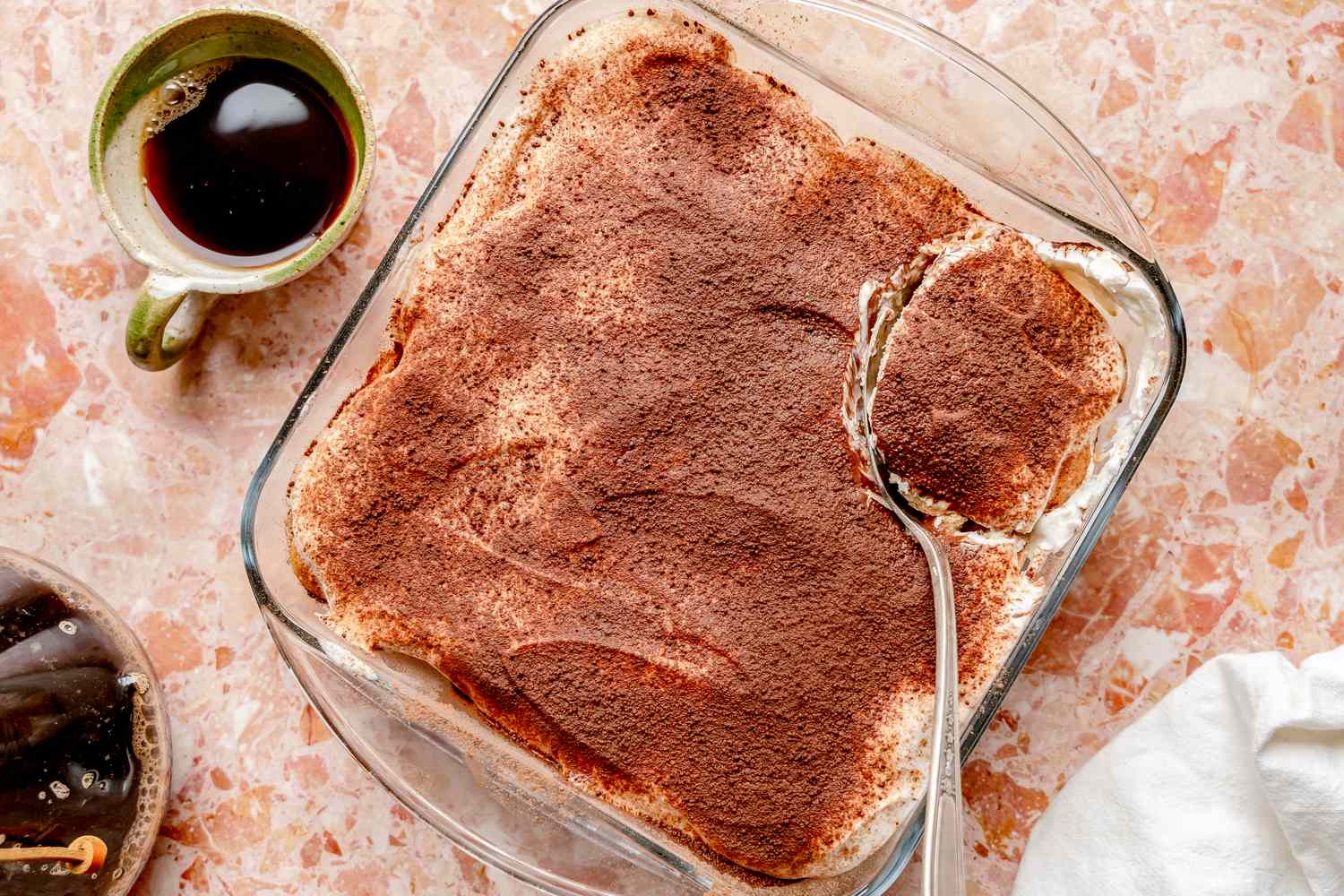 A finished tiramisu in a glass baking dish topped with a thick dusting of cocoa powder, with a spoon scoop showing creamy layers and coffee-soaked ladyfingers on a bright kitchen counter