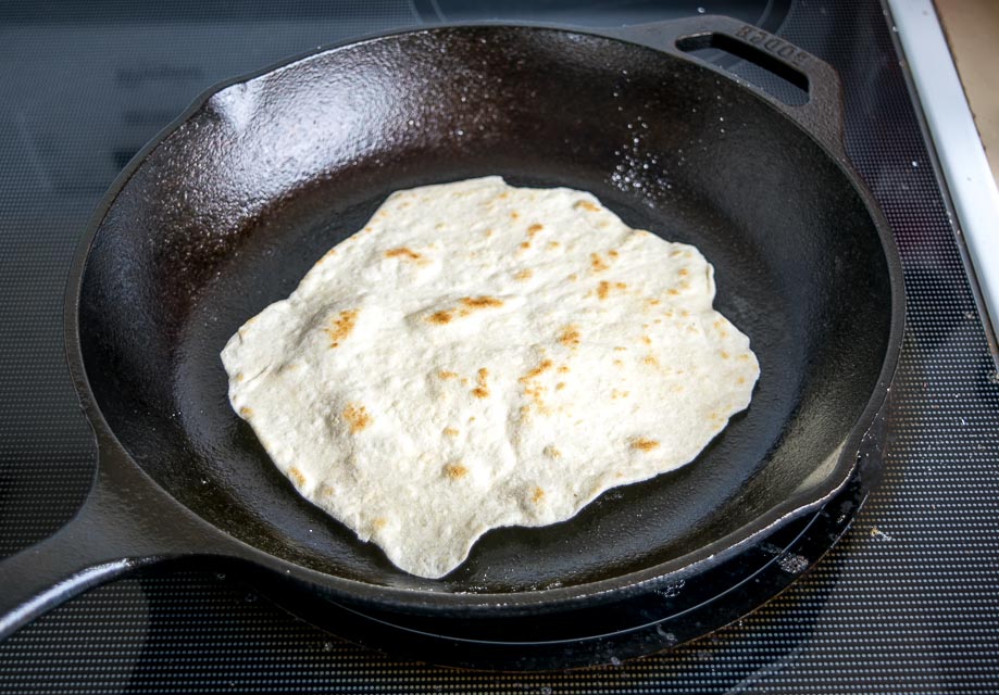 A flour tortilla being placed into a lightly oiled skillet on a stovetop, warm kitchen lighting