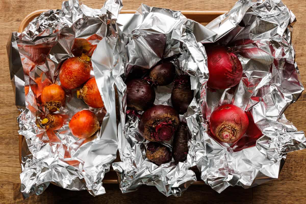 A foil packet opened on a sheet pan revealing roasted beets with their skins loosened, sitting on a neutral-toned kitchen counter