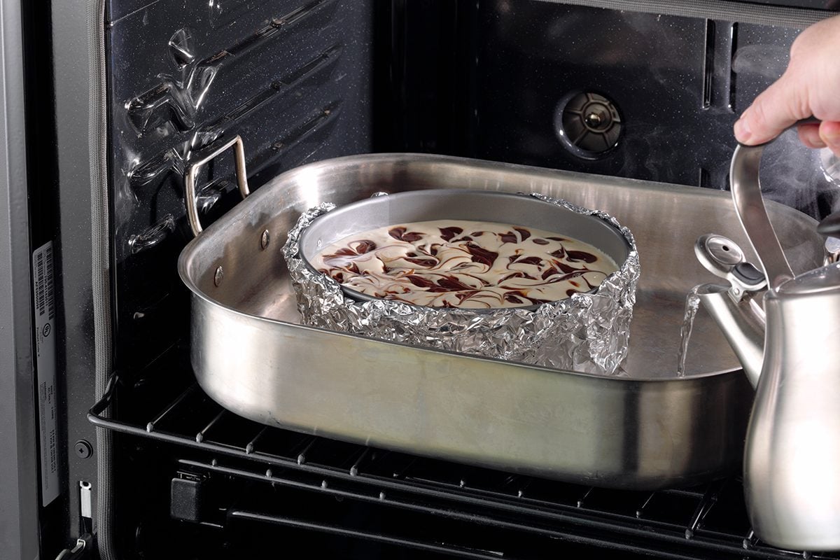 A foil-wrapped springform pan sitting inside a larger roasting pan with hot water around it on a kitchen counter