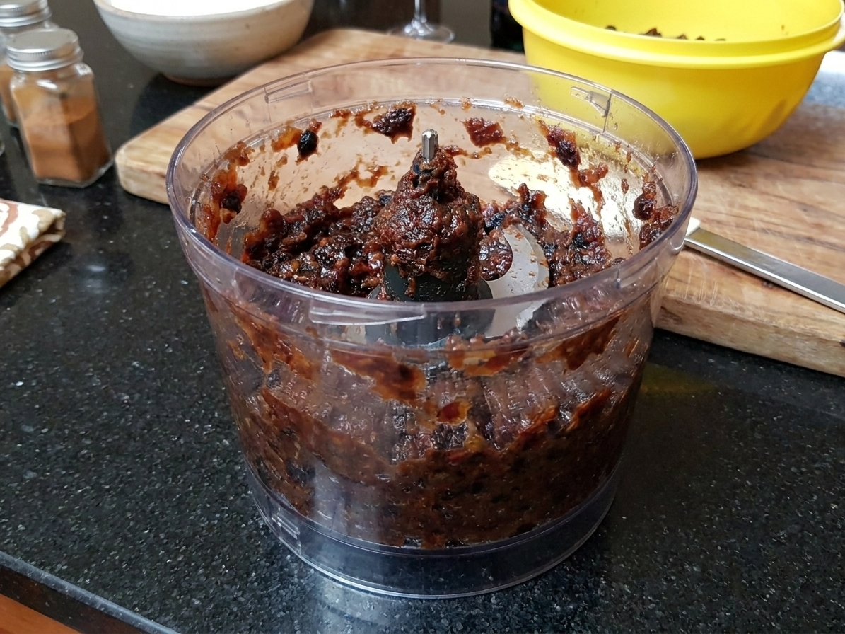 A food processor bowl filled with dark, thick blended soaked fruit paste for black cake