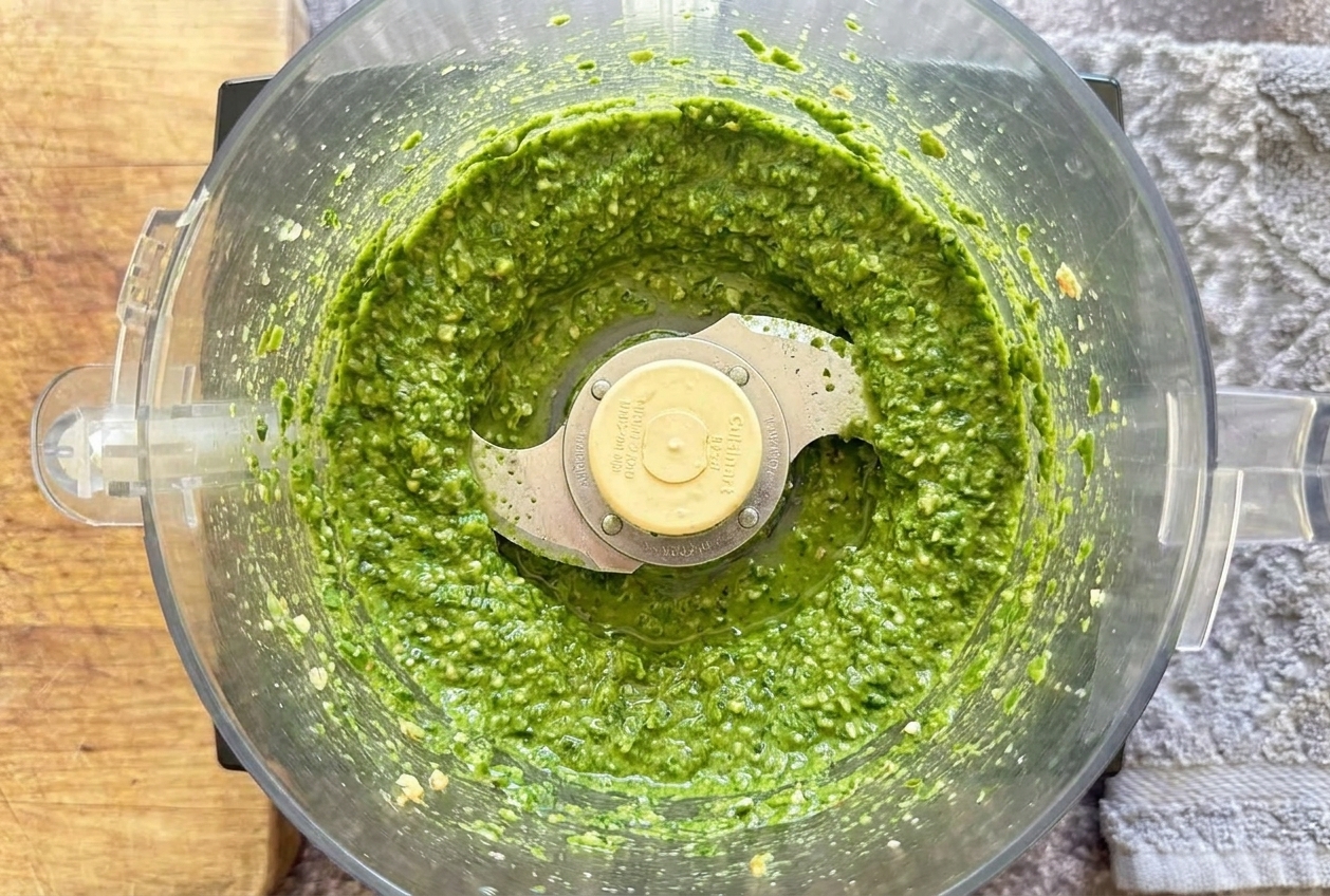 A food processor bowl with bright green garlic scape pesto mid-blend, with pesto clinging to the sides