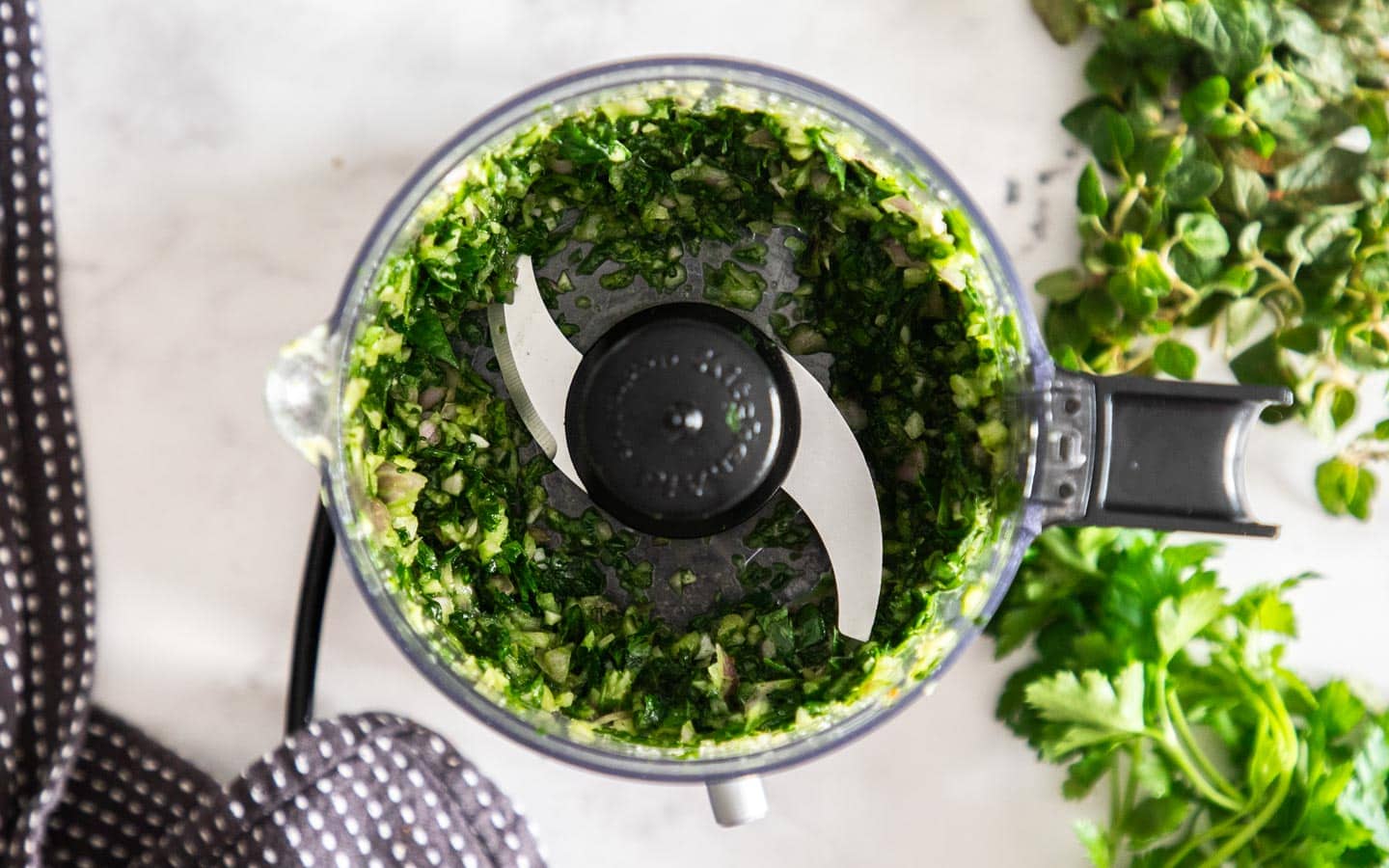 A food processor with red chimichurri in progress, with visible herbs and red pepper pieces on a home kitchen counter