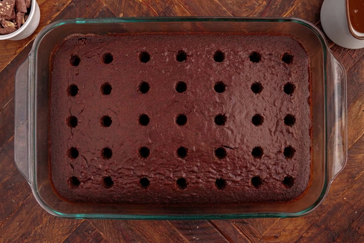 A fresh baked chocolate sheet cake in a 9x13 pan with evenly spaced poke holes across the surface, shot on a warm kitchen counter