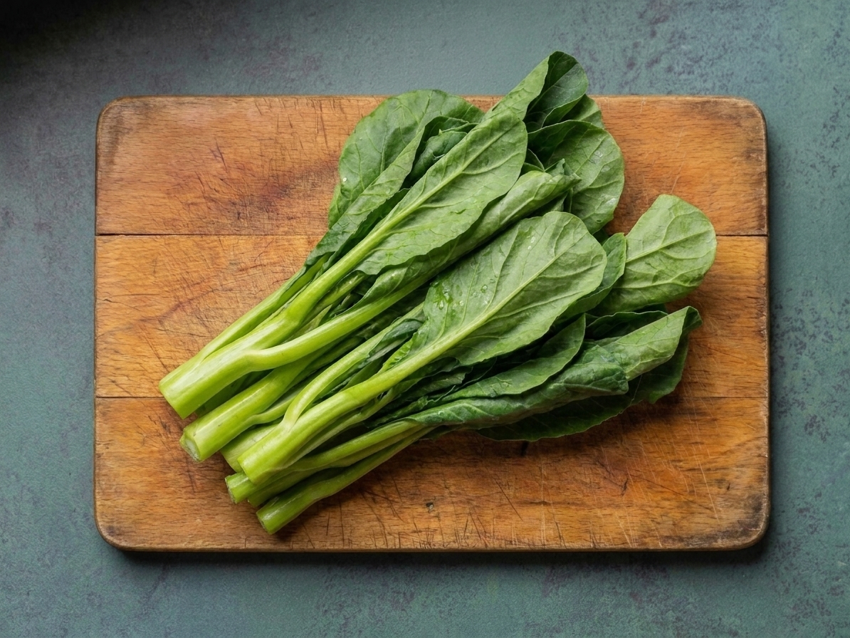 A fresh bunch of raw gai lan (Chinese broccoli) with thick stems and dark green leaves on a wooden cutting board on a bright kitchen counter