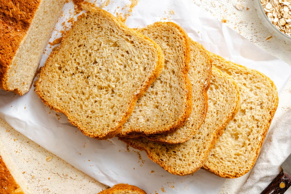 A fresh honey oat sandwich bread loaf resting on a wooden cutting board beside a Cuisinart bread maker on a bright kitchen counter