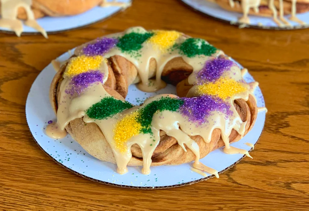 A freshly baked Mardi Gras king cake shaped in a ring, glazed and topped with purple, green, and gold sanding sugar on a wooden table
