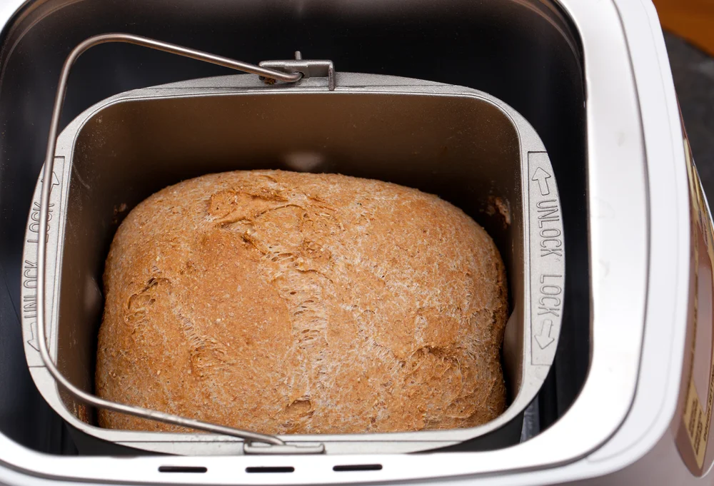 A freshly baked gluten free sandwich bread loaf sliced on a wooden cutting board beside a bread machine on a bright kitchen counter