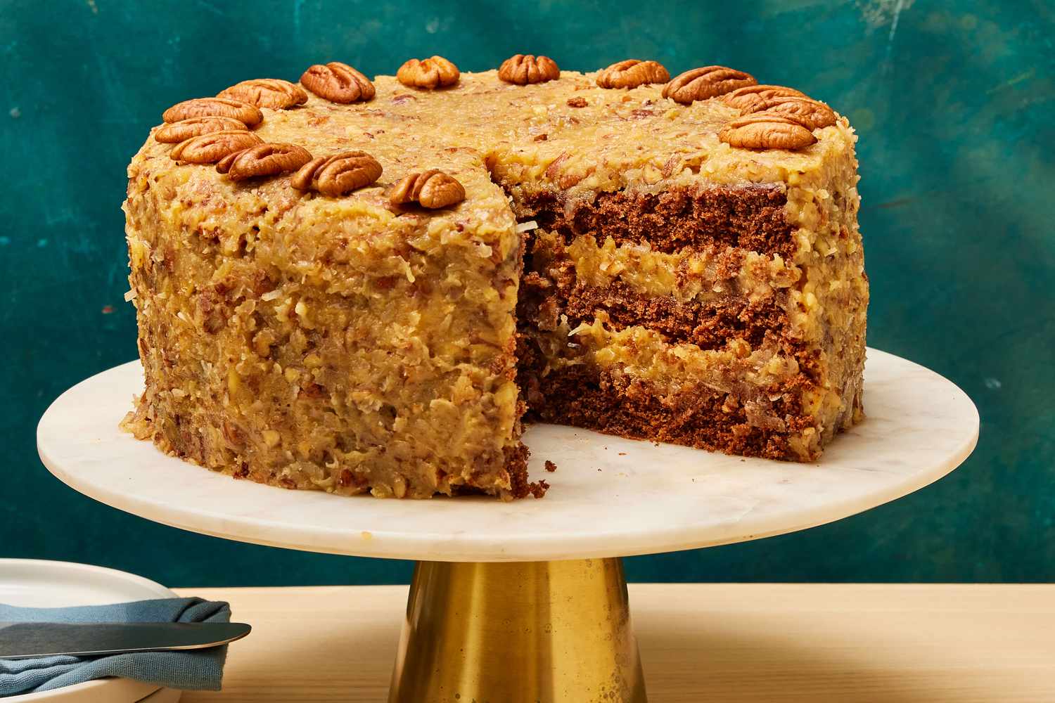 A freshly frosted German chocolate cake on a simple cake stand, topped with thick coconut-pecan frosting that looks glossy and caramel-brown in warm kitchen light