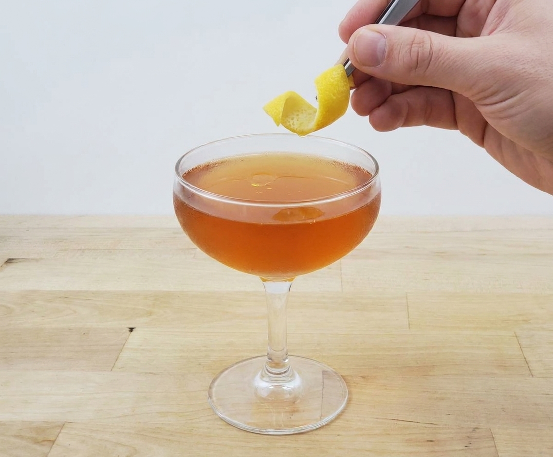A freshly strained Paper Plane cocktail in a coupe glass as a lemon twist is held above it, capturing the moment before serving