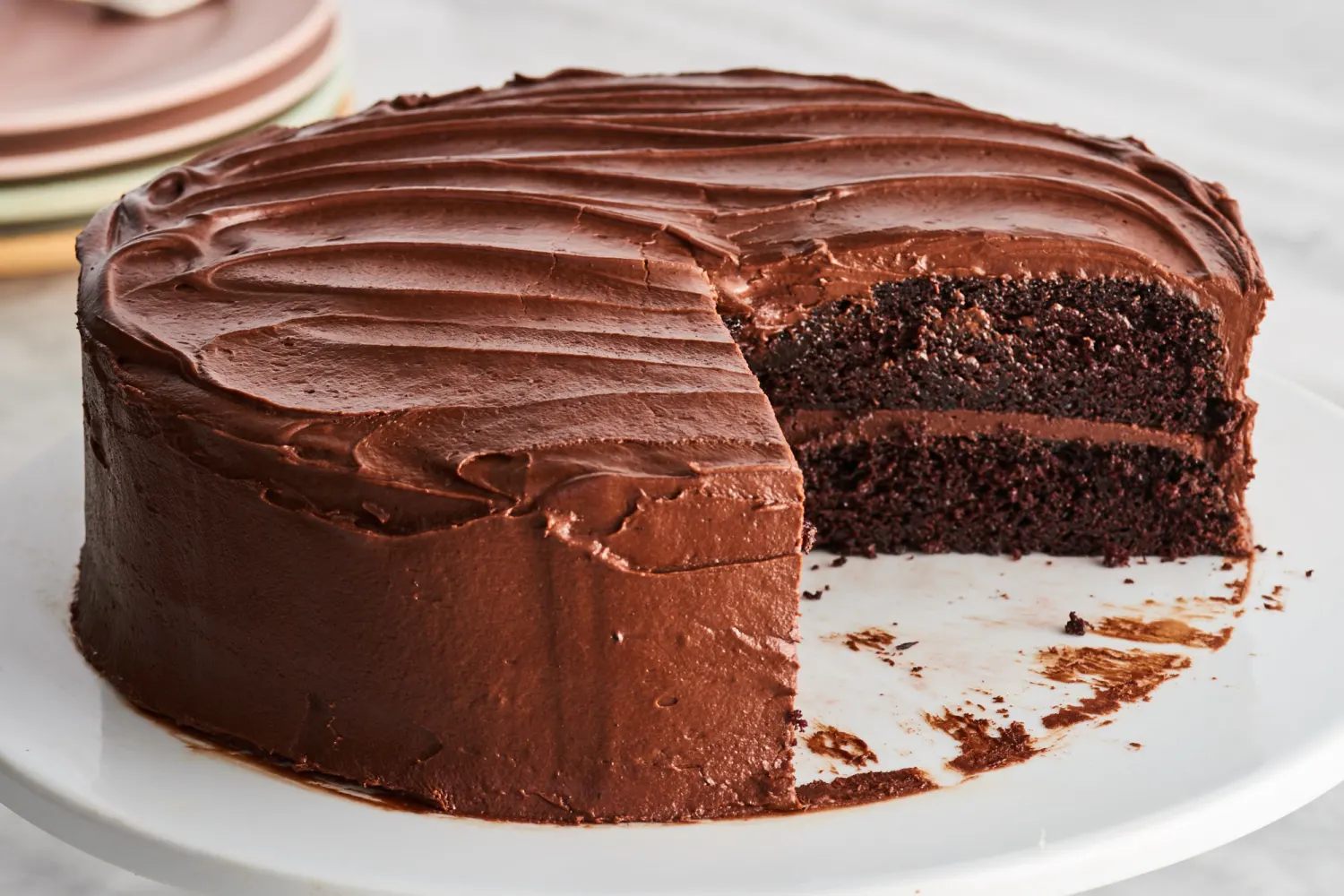 A frosted chocolate layer cake on a simple cake stand on a bright kitchen counter, with one slice cut to show the moist crumb