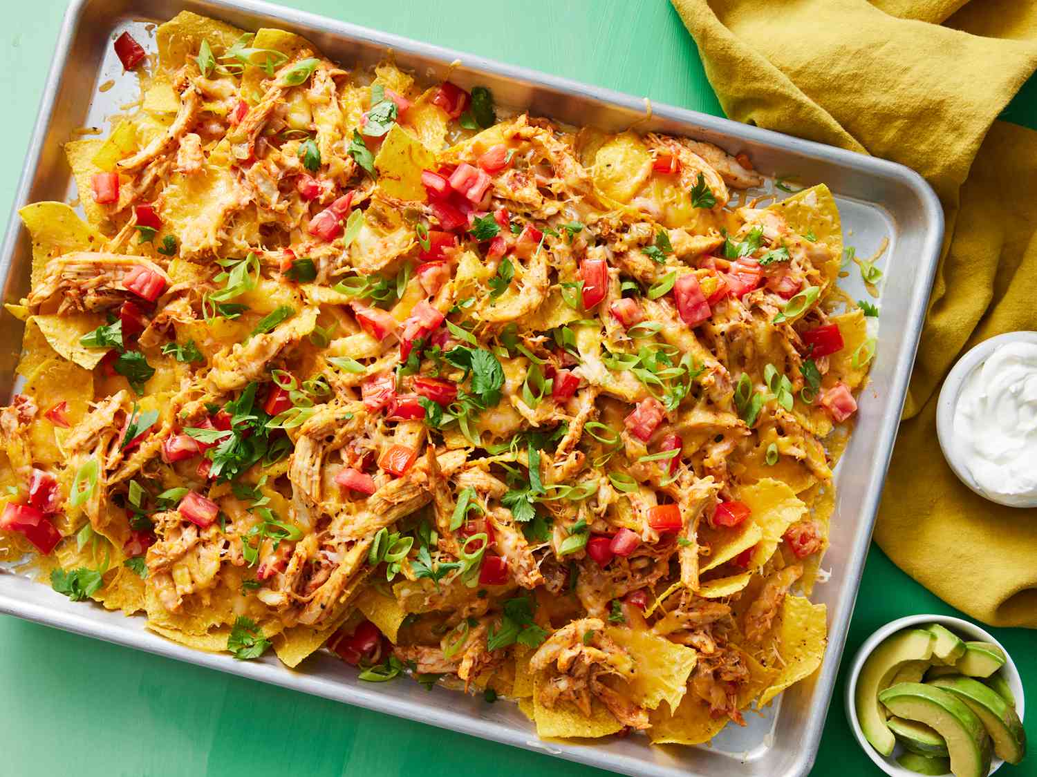A full sheet pan of chicken nachos with melted cheddar and Monterey Jack, black beans, jalapeños, cilantro, and dollops of lime crema on a kitchen counter under warm lighting