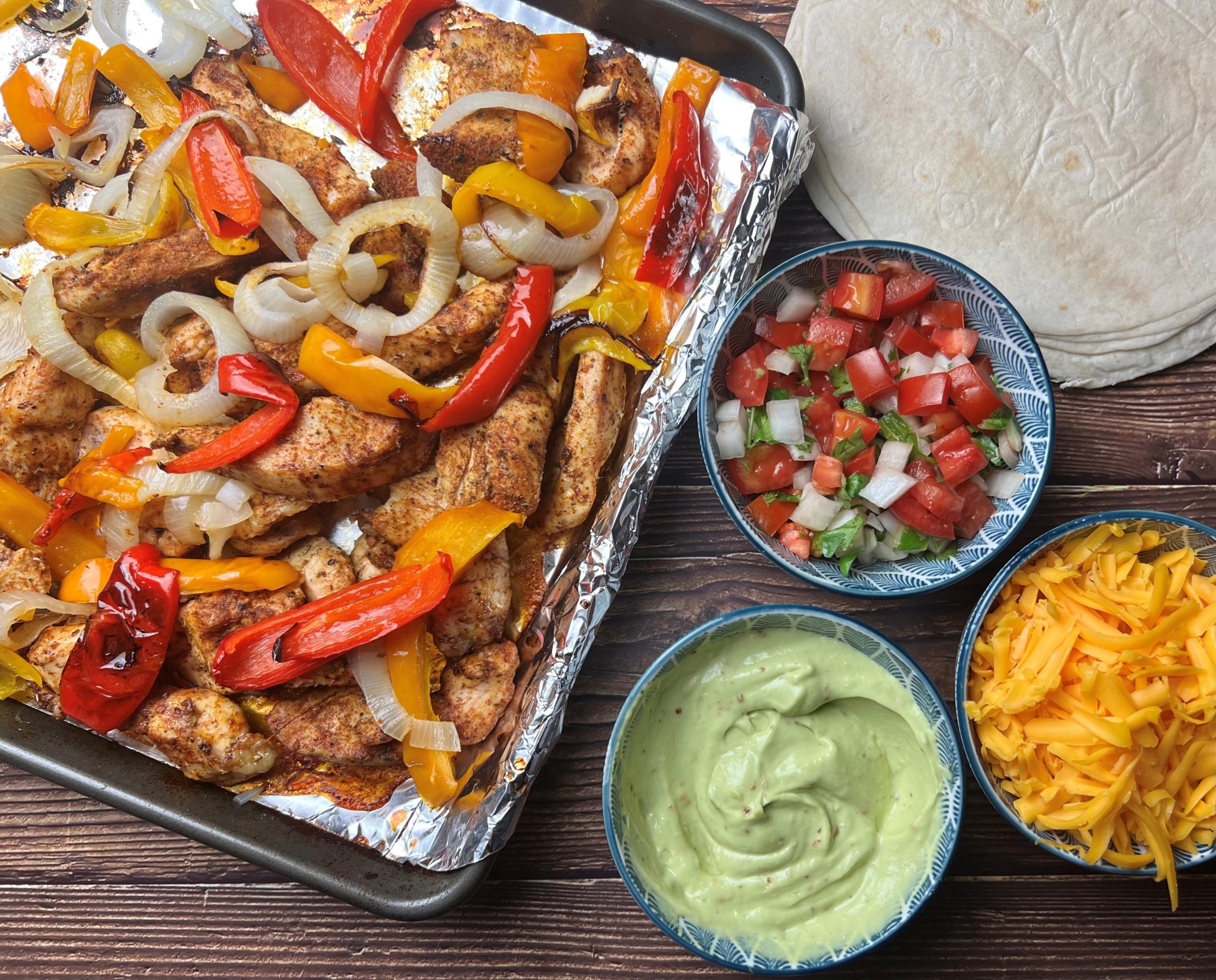 A full sheet pan of roasted chicken strips with sliced bell peppers and onions, lightly charred at the edges, with lime wedges on the side on a kitchen counter