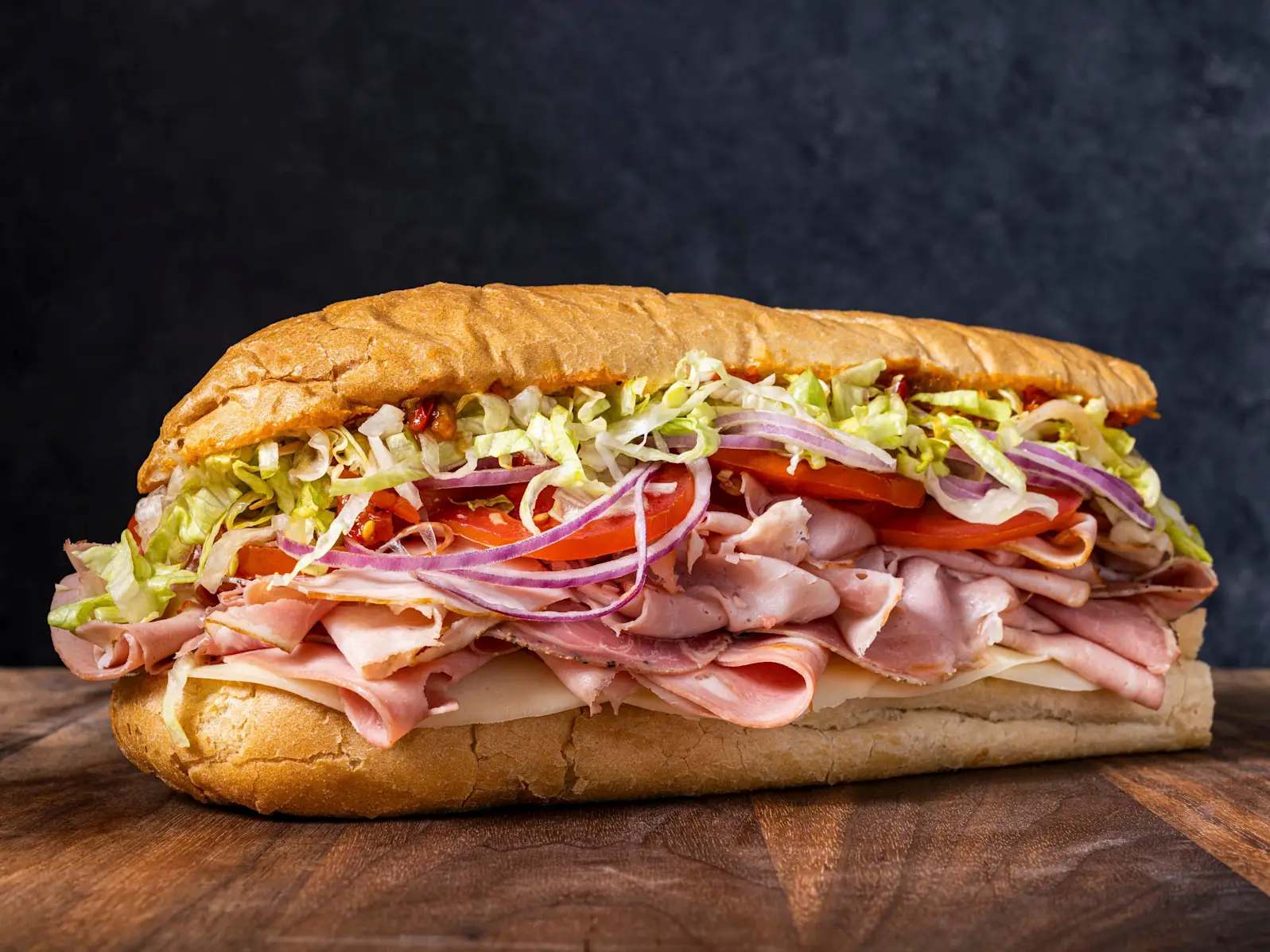 A fully assembled Italian sub sandwich cut into halves on a wooden cutting board, with shredded lettuce, tomato, onion, provolone, and layered cured meats visible in warm kitchen lighting
