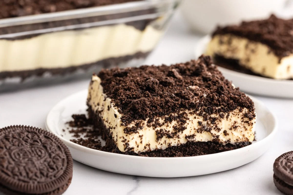 A glass 9x13-inch baking dish filled with Oreo dirt cake, showing creamy chocolate pudding layers and a thick crumb topping, on a bright kitchen counter with a serving spoon