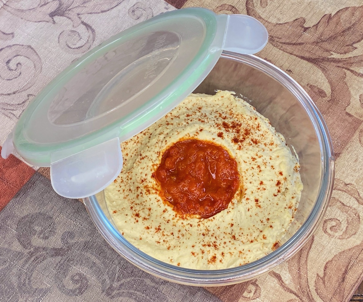 A glass airtight container filled with hummus, topped with a thin layer of olive oil