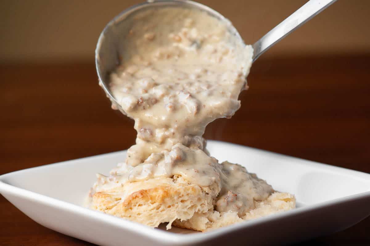 A glass airtight container filled with leftover sausage gravy cooling on a kitchen counter next to a small saucepan and spoon