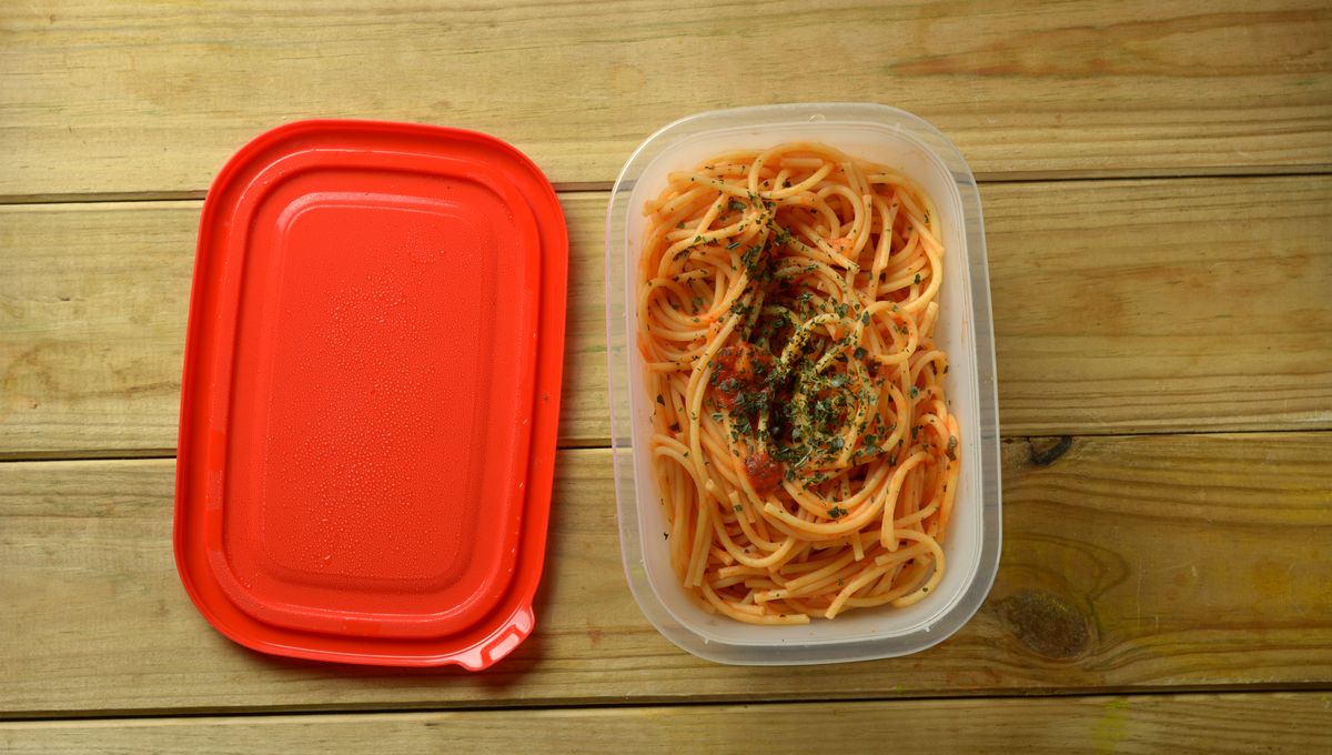 A glass airtight container holding leftover linguine with clam sauce on a kitchen counter next to a small spoon and a lemon wedge