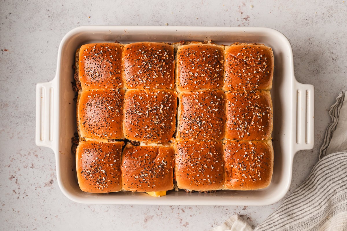 A glass baking dish filled with assembled cheeseburger sliders before baking, showing seasoned beef and cheddar cheese layered inside soft rolls on a kitchen counter