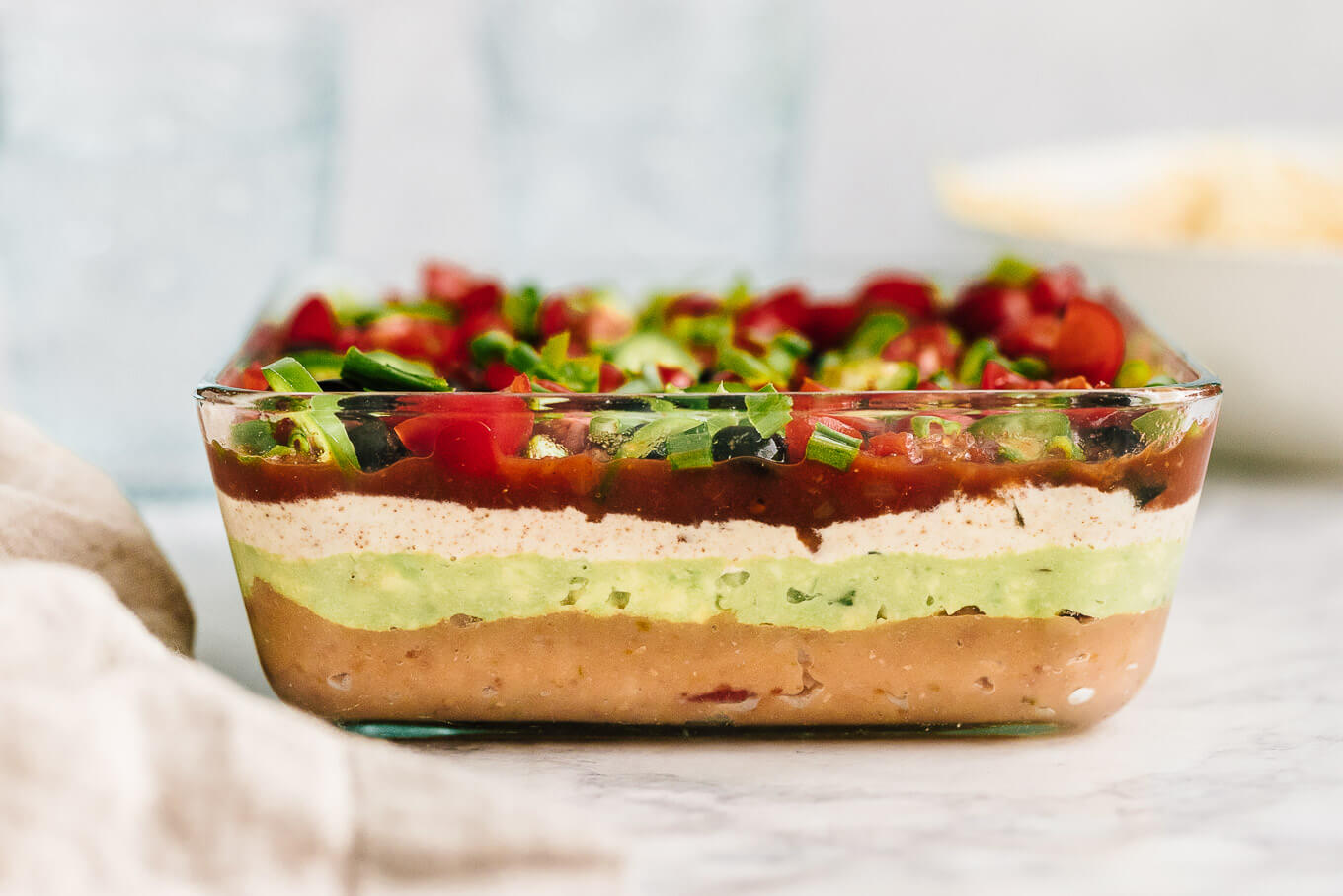 A glass baking dish filled with seven layer taco dip topped with shredded cheese, diced tomatoes, olives, and green onions on a bright kitchen counter with tortilla chips nearby