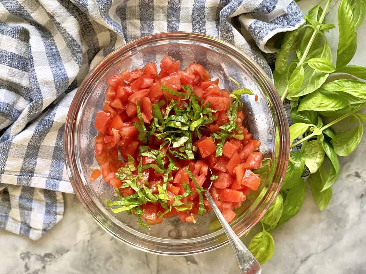 A glass bowl filled with diced tomatoes, sliced basil, and olive oil being stirred with a spoon on a kitchen counter