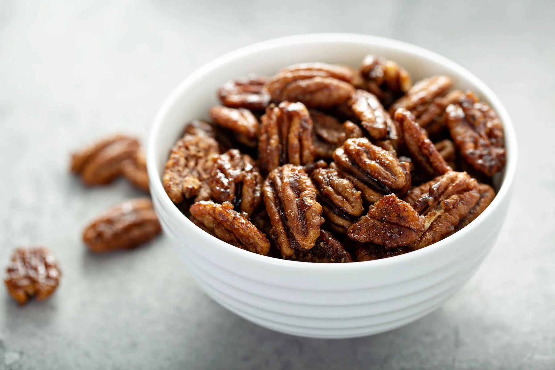 A glass bowl filled with glossy praline pecans on a light kitchen counter with a linen napkin nearby
