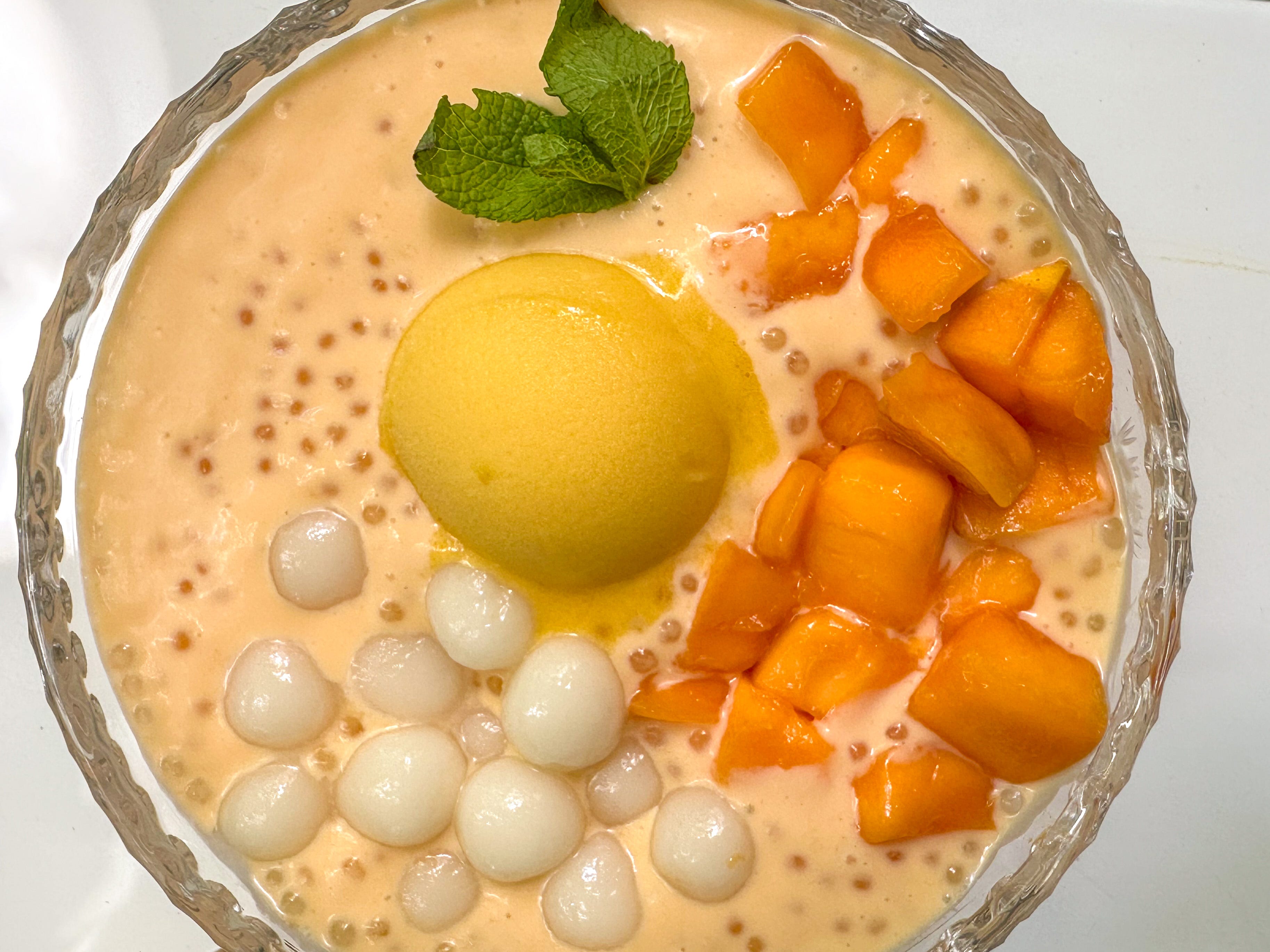 A glass bowl of creamy mango sago topped with fresh mango cubes and a few tapioca pearls, set on a light kitchen table with soft natural light