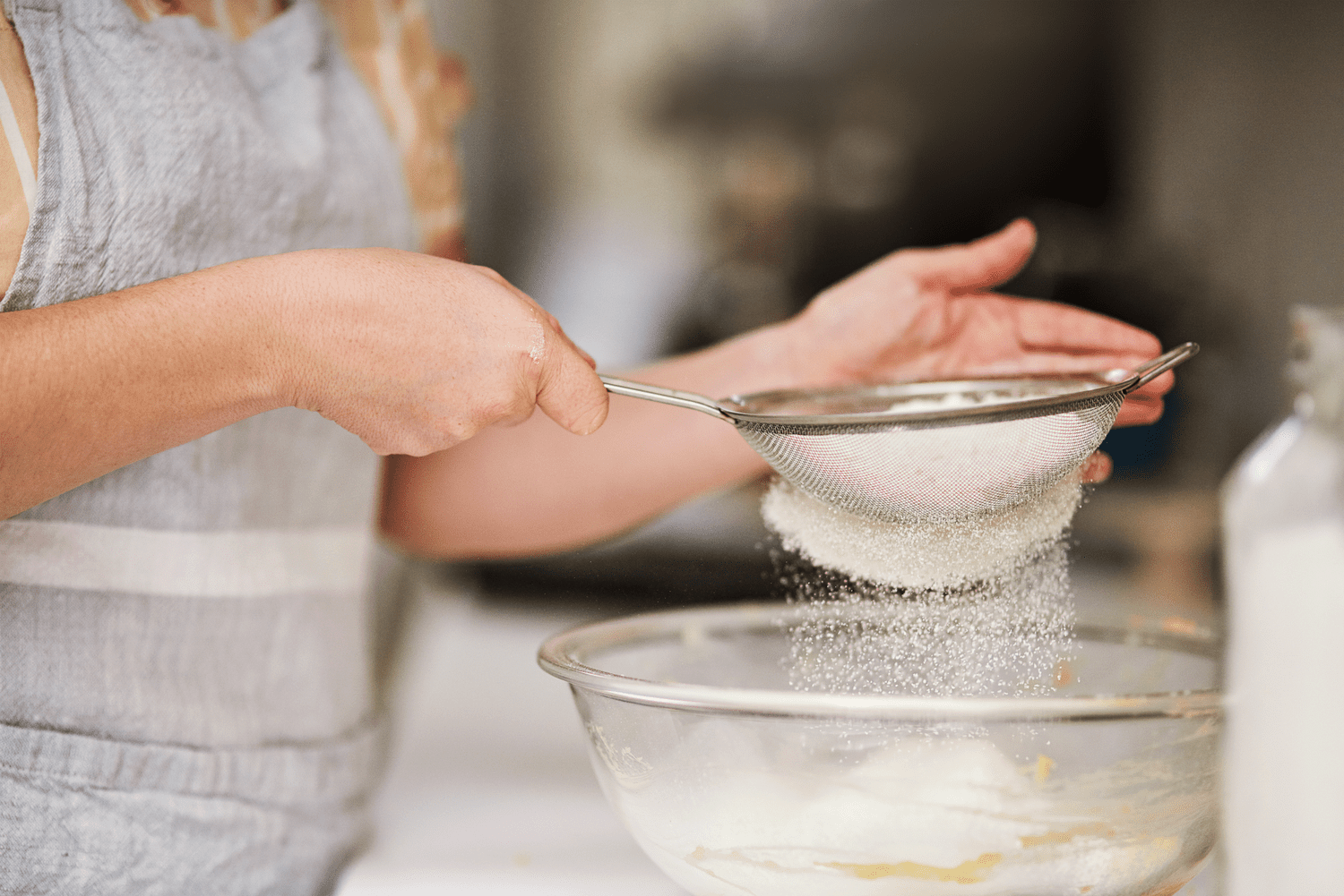 A glass bowl of freshly sifted powdered sugar on a kitchen counter next to a fine mesh sieve