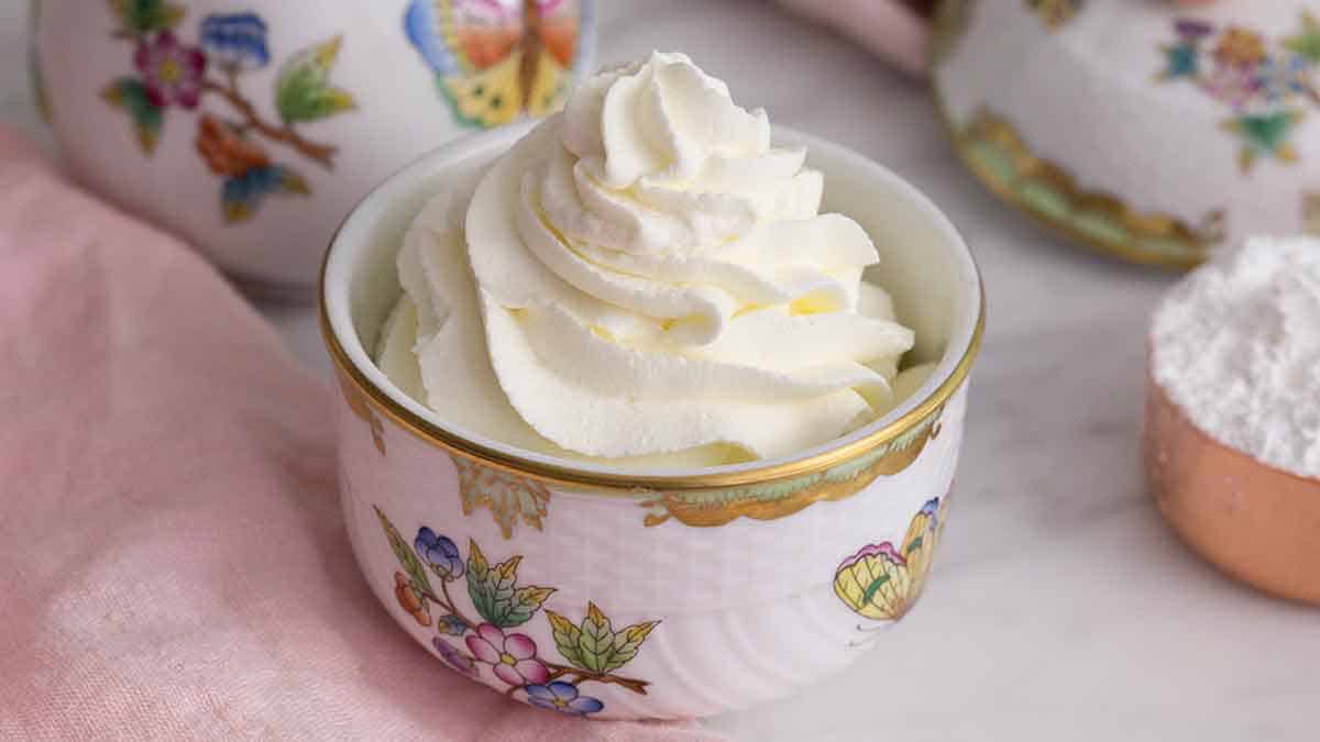 A glass bowl of freshly whipped cream with soft, glossy peaks on a light-colored kitchen counter with a whisk nearby