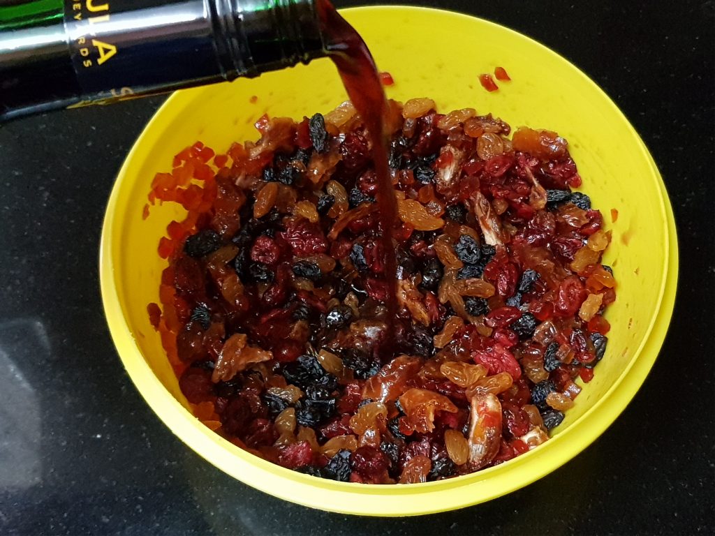 A glass bowl of mixed dried fruit soaking in orange juice with a spoon resting inside on a kitchen counter