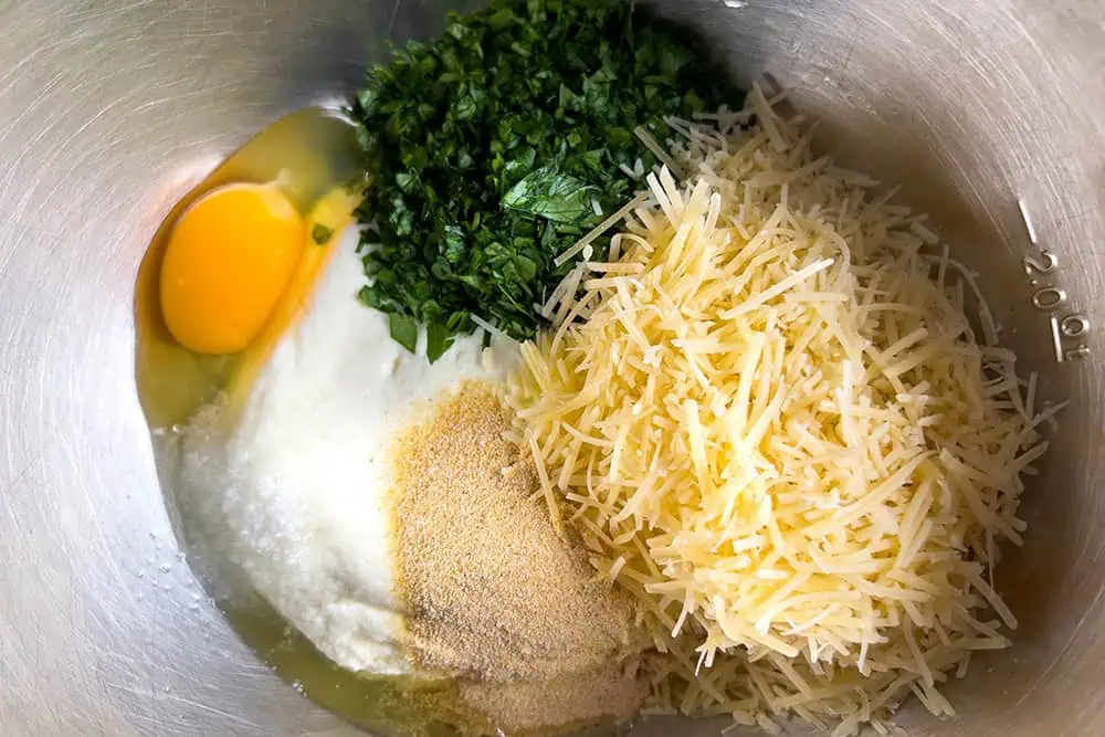 A glass bowl on a kitchen counter with ricotta, egg, and Parmesan being mixed with a spoon