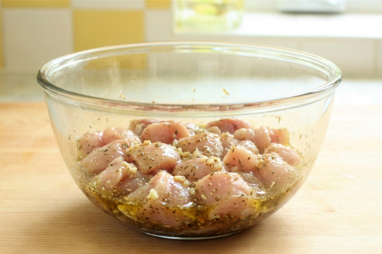 A glass bowl with chicken pieces coated in lemony olive oil marinade with visible oregano and minced garlic on a kitchen counter