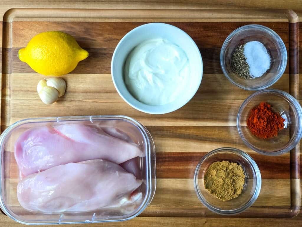 A glass bowl with lemon juice, olive oil, minced garlic, and spices measured out on a kitchen counter with a lemon half nearby