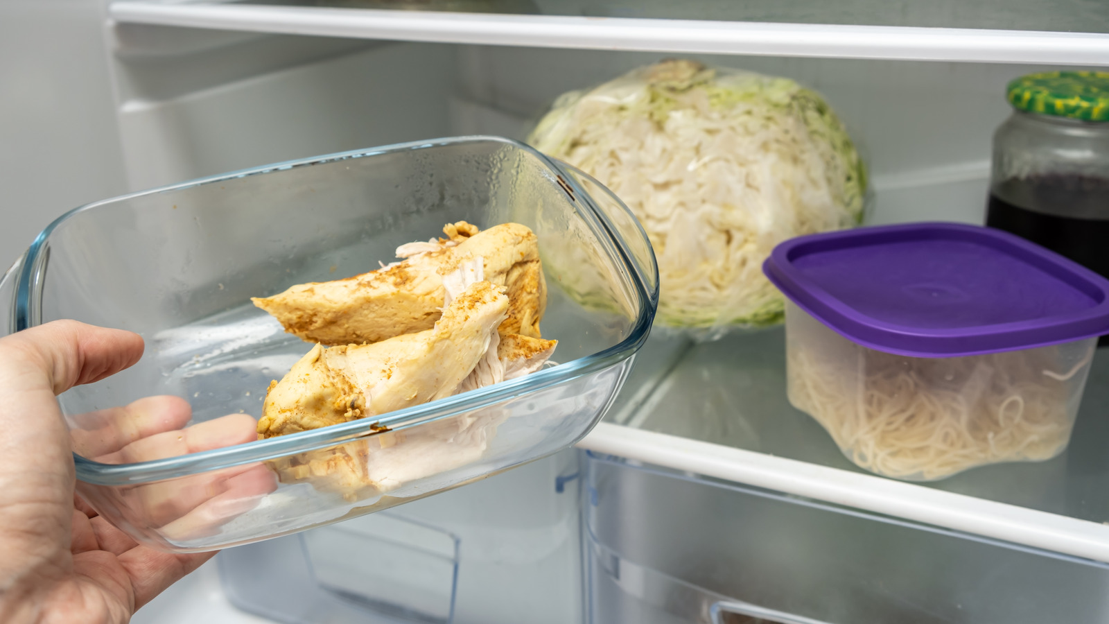A glass container of sliced cooked chicken shawarma cooling on a kitchen counter next to a lidded container of yogurt sauce