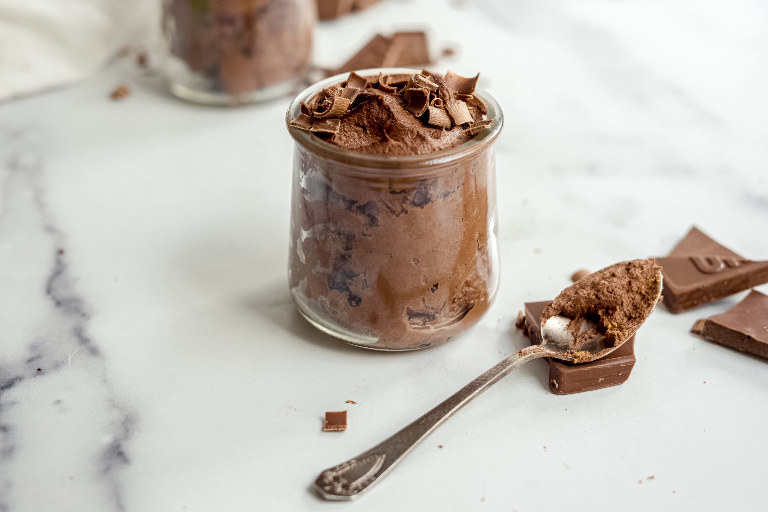 A glass cup filled with thick chocolate protein pudding topped with raspberries and chocolate shavings on a light kitchen counter with a spoon beside it