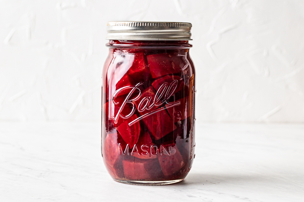 A glass jar filled with deep ruby pickled beet slices in brine on a light kitchen counter, with a few peppercorns and bay leaf visible