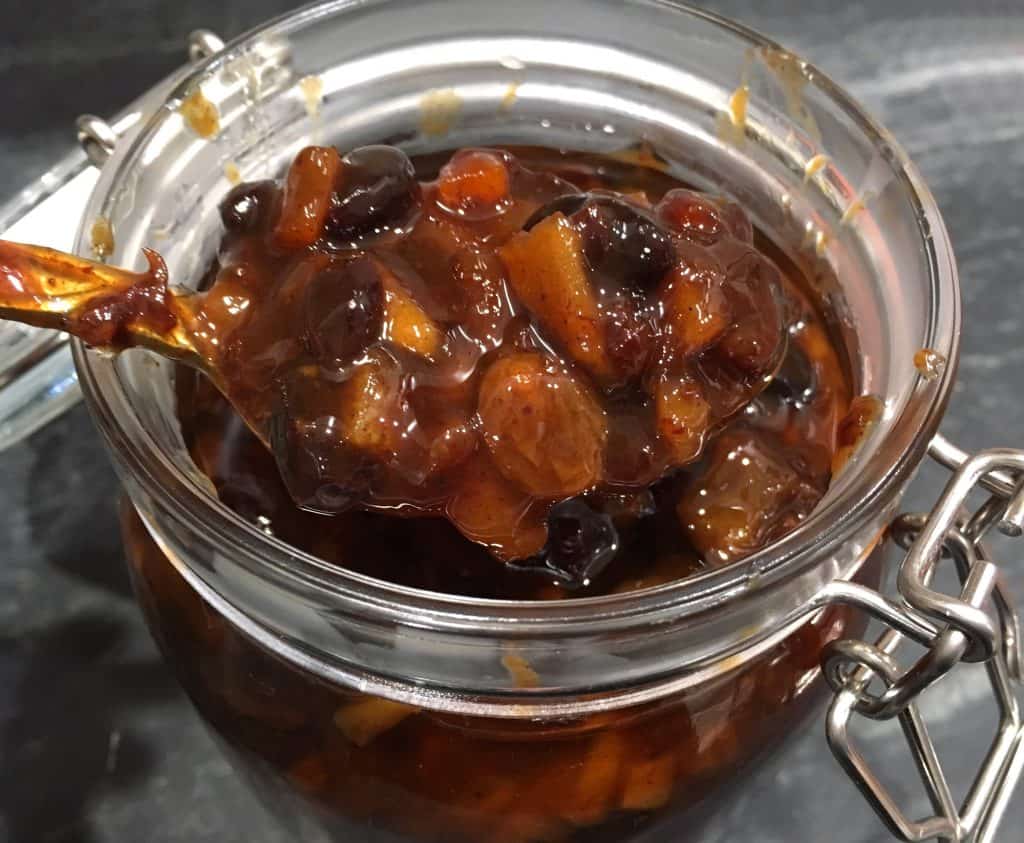 A glass jar filled with glossy homemade mincemeat, with visible raisins and orange zest, sitting on a warm-toned kitchen counter beside a wooden spoon