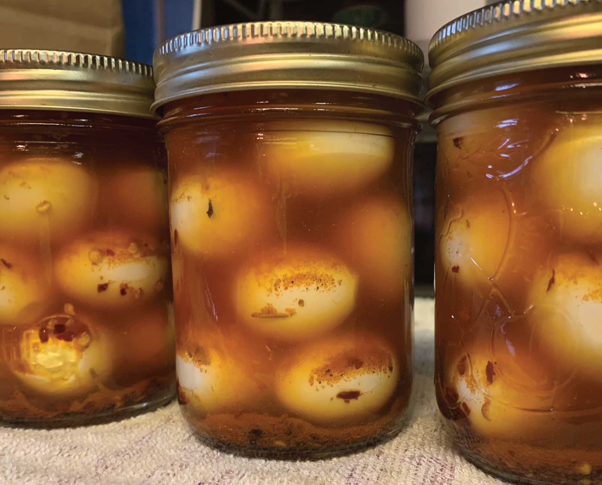 A glass jar in a refrigerator holding peeled quail eggs submerged in a dark soy marinade with sliced green onions