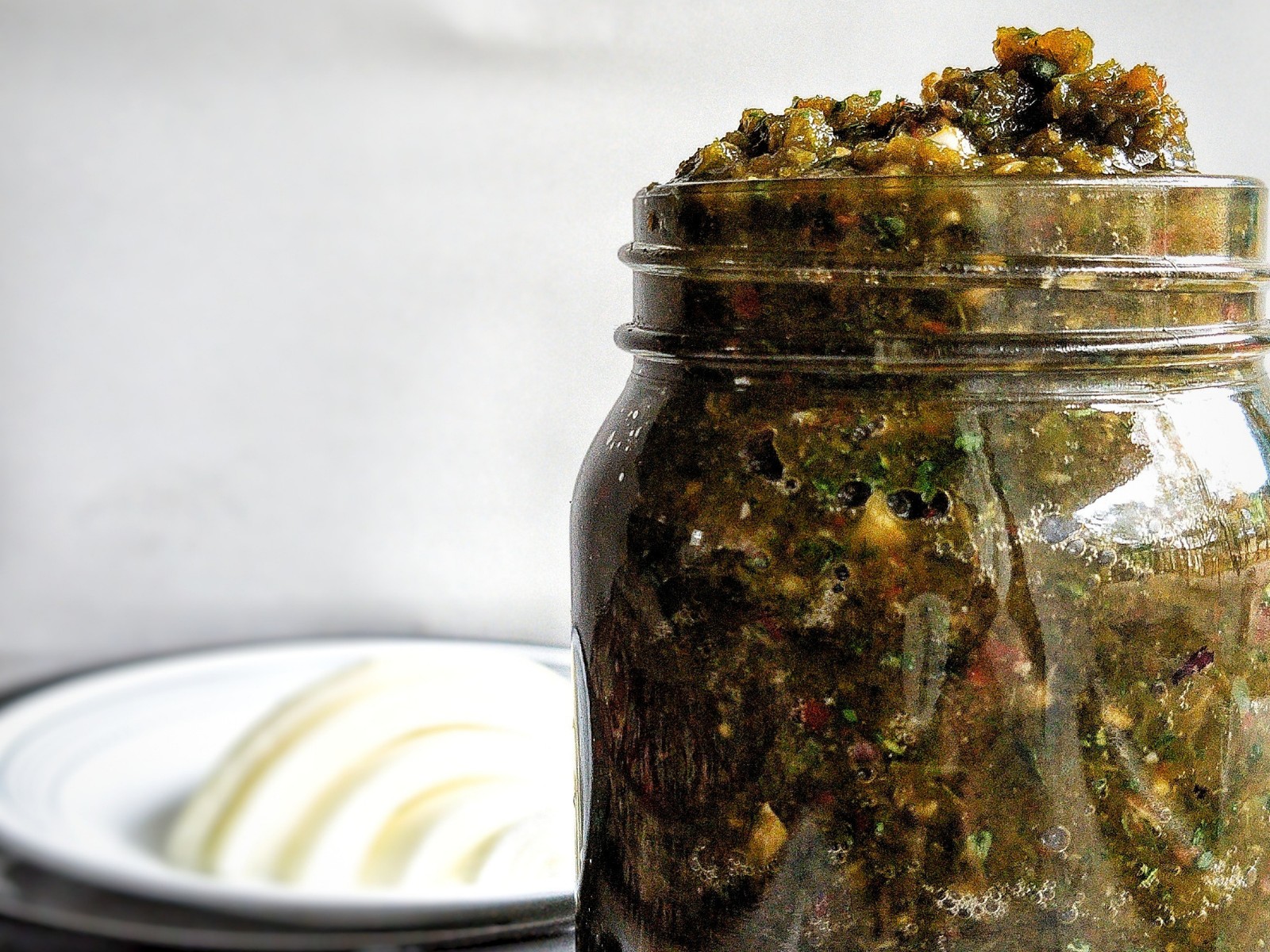 A glass jar of bright green Puerto Rican sofrito on a sunlit kitchen counter with cilantro, garlic, onion, and green peppers nearby