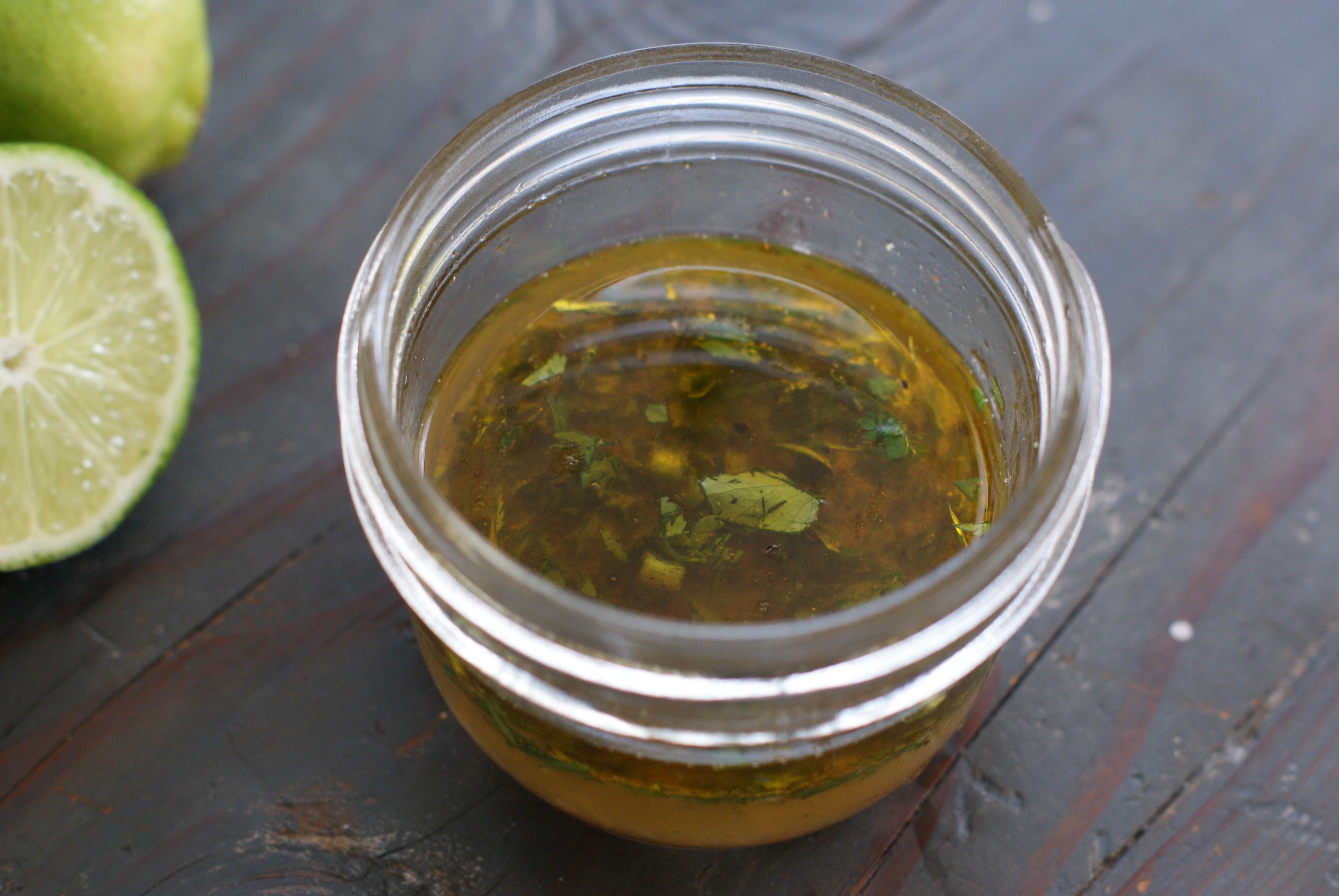 A glass jar of creamy chipotle vinaigrette with visible flecks of red chipotle and spices, sitting in a bright kitchen beside a halved lime and a small bowl of salad