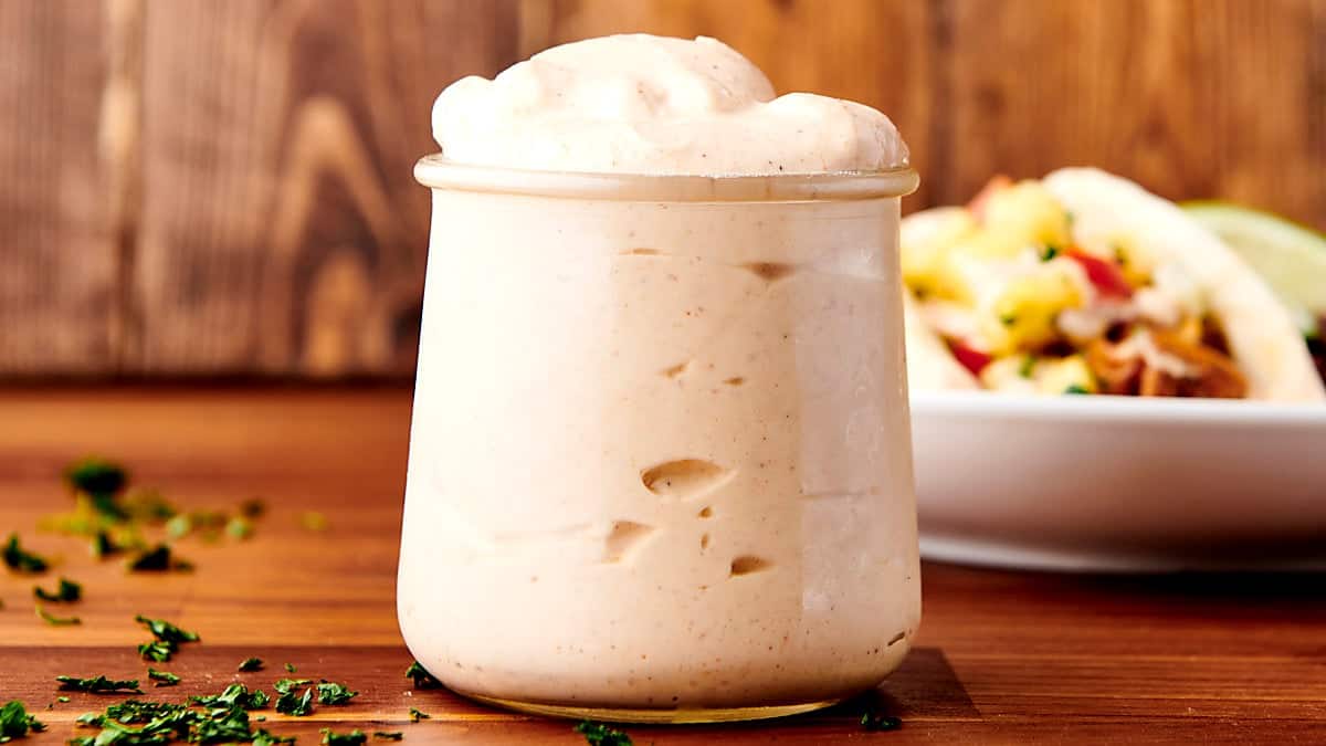 A glass jar of creamy fish taco sauce with a lid beside it on a kitchen counter, with a small spoon and lime wedge nearby